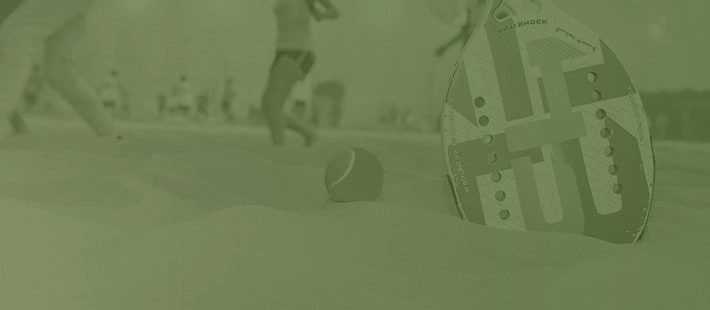 Raquete te beach tennis na areia ao lado de bolinha - ao fundo pessoas praticando esportes.